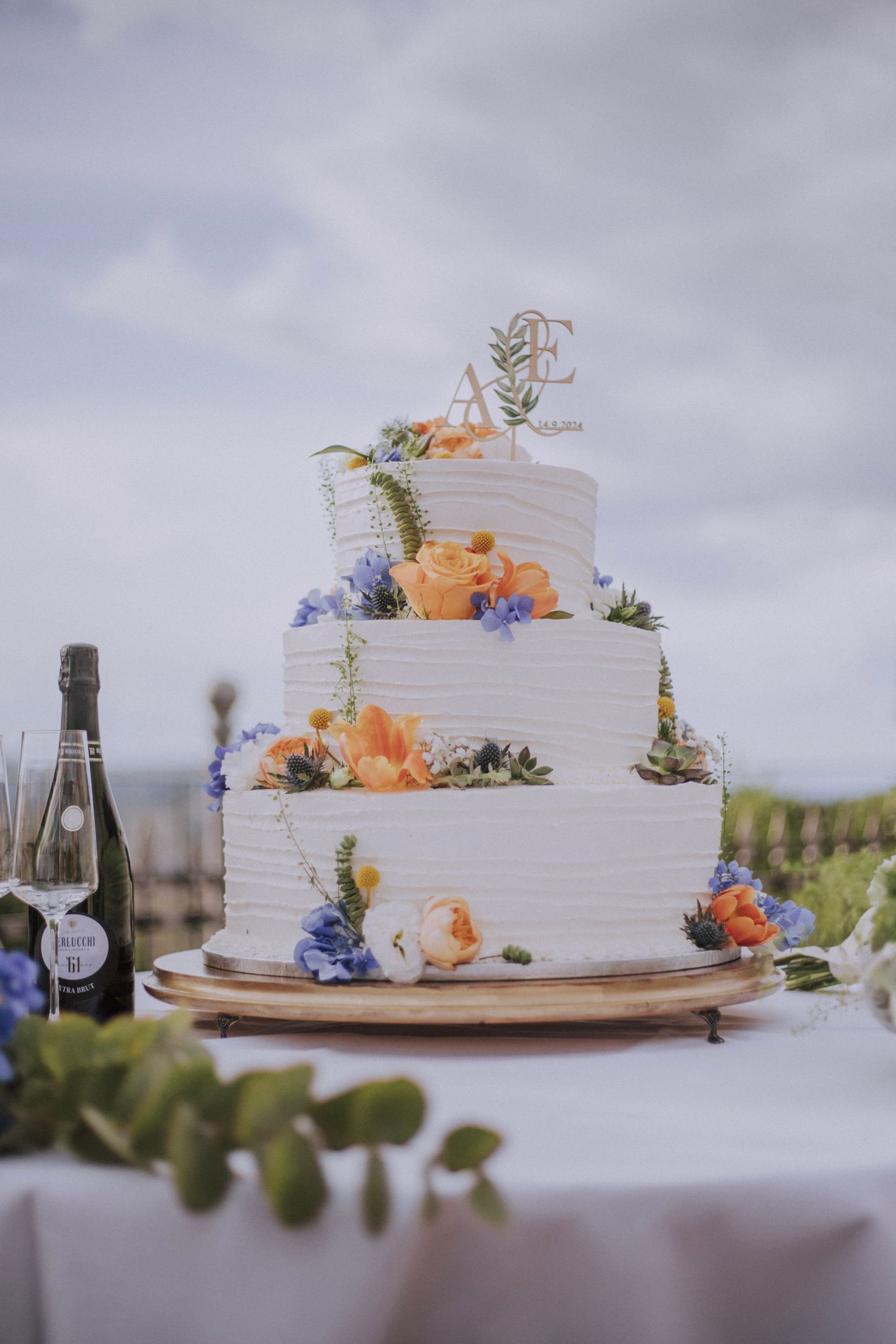 An elegant three-tier white wedding cake decorated with peach and blue flowers set against a cloudy sky.