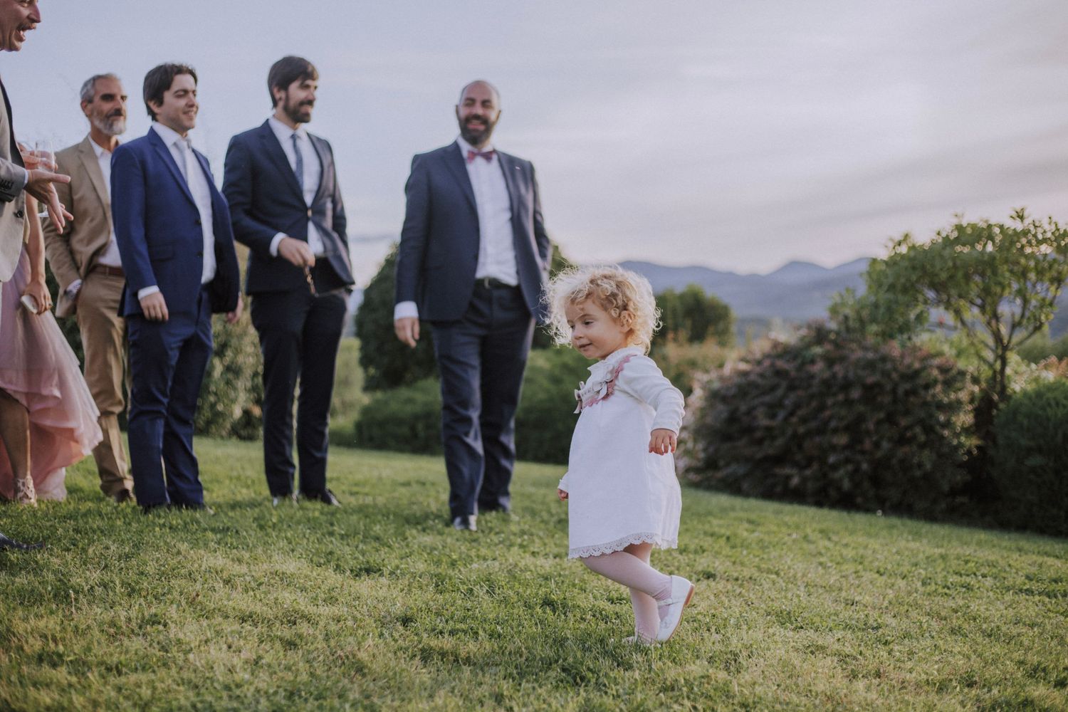 A candid wedding celebration moment on a grassy hilltop with guests and a small child.