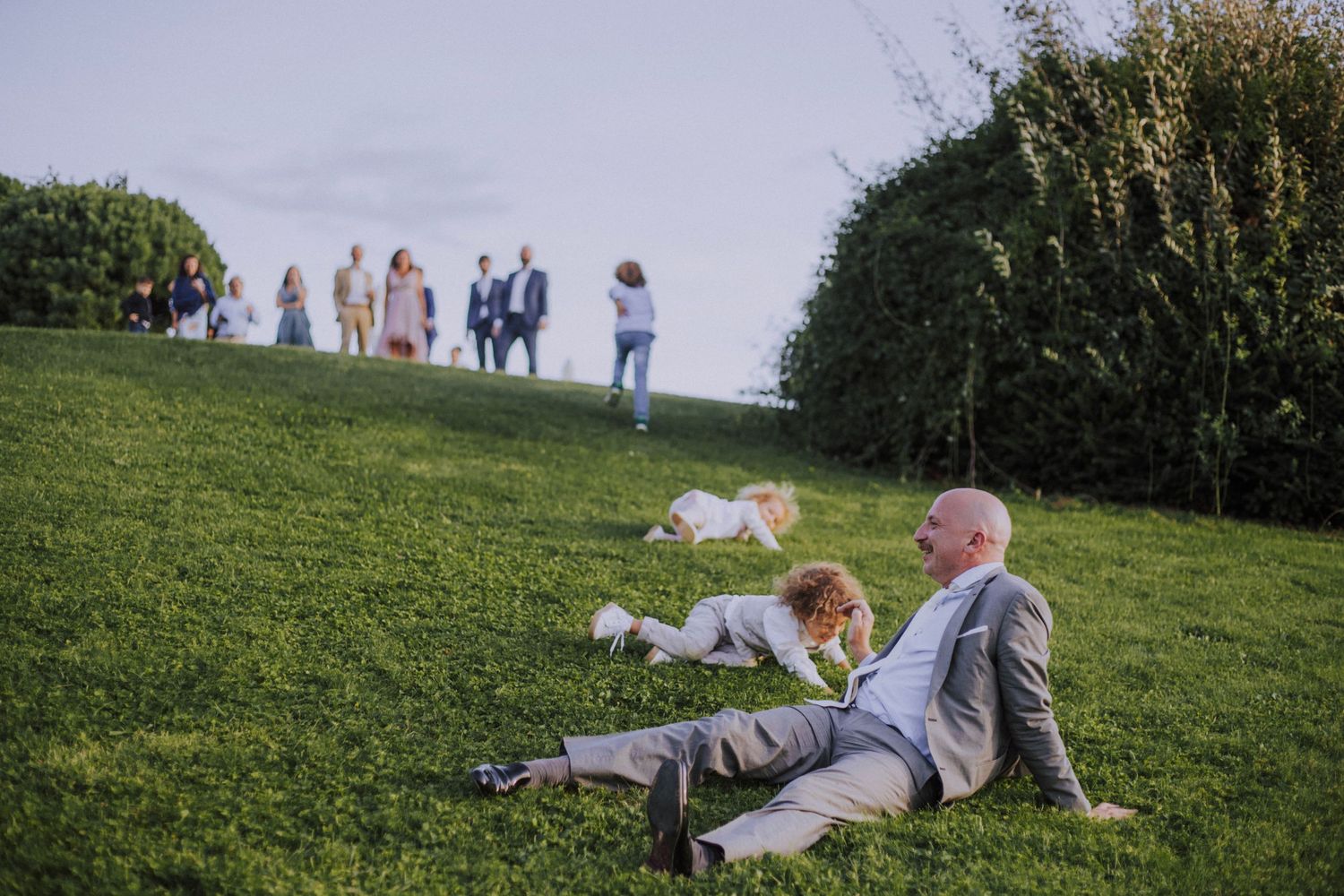 A relaxed guest resting on grass while others socialize during an outdoor wedding reception.