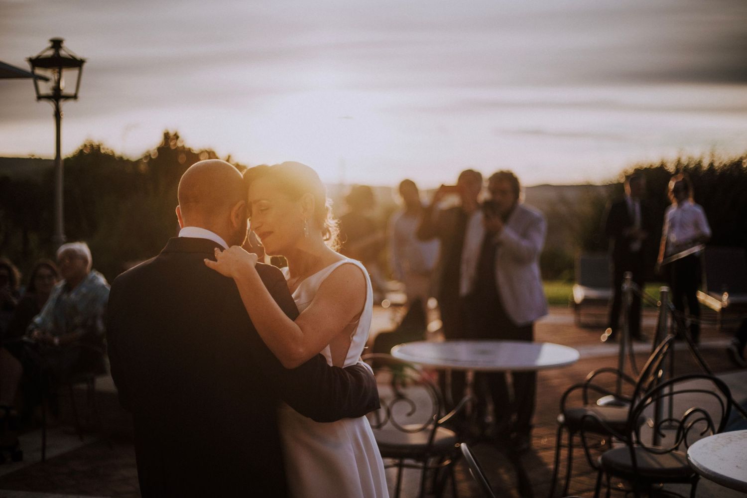 Couple shares a romantic dance at sunset while wedding guests look on in the background.