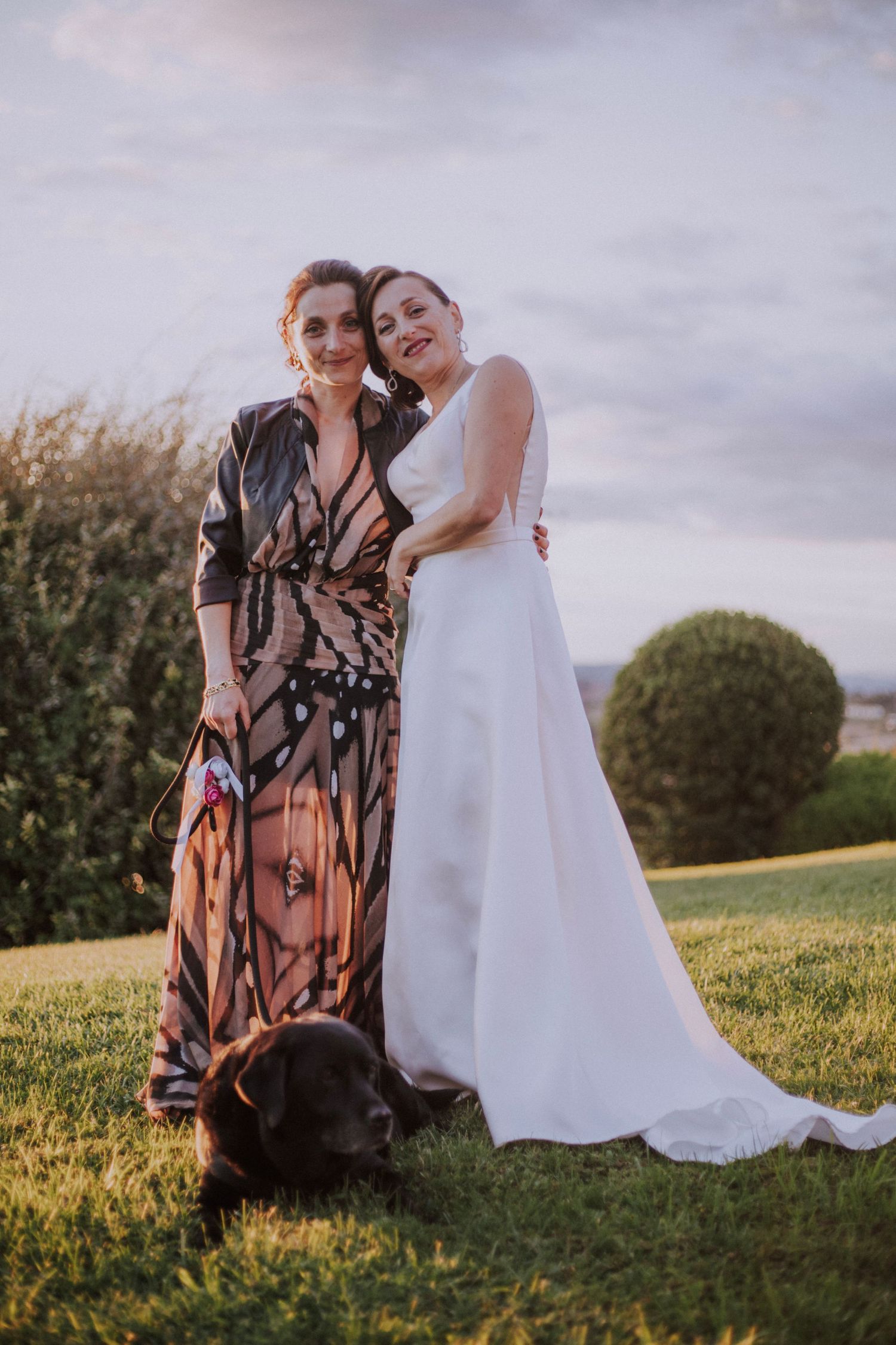 Bride in white gown poses with guest in patterned dress on grassy hill with black dog.