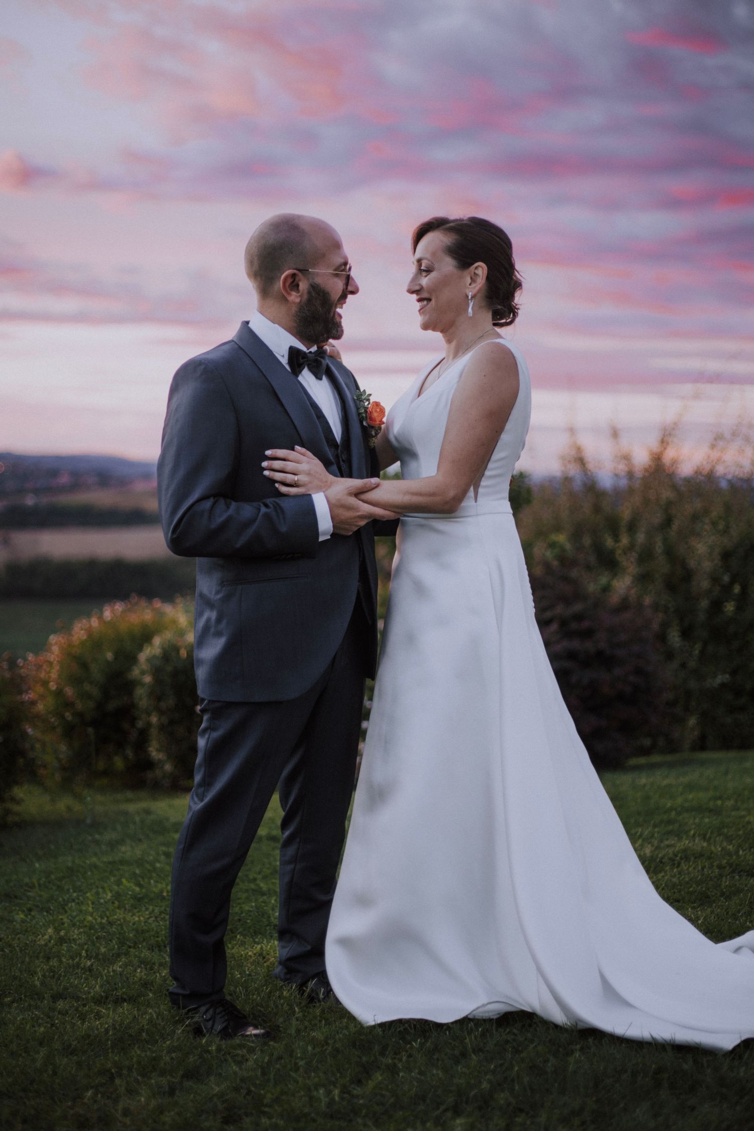 Couple shares intimate moment against stunning pink sunset sky during outdoor wedding.