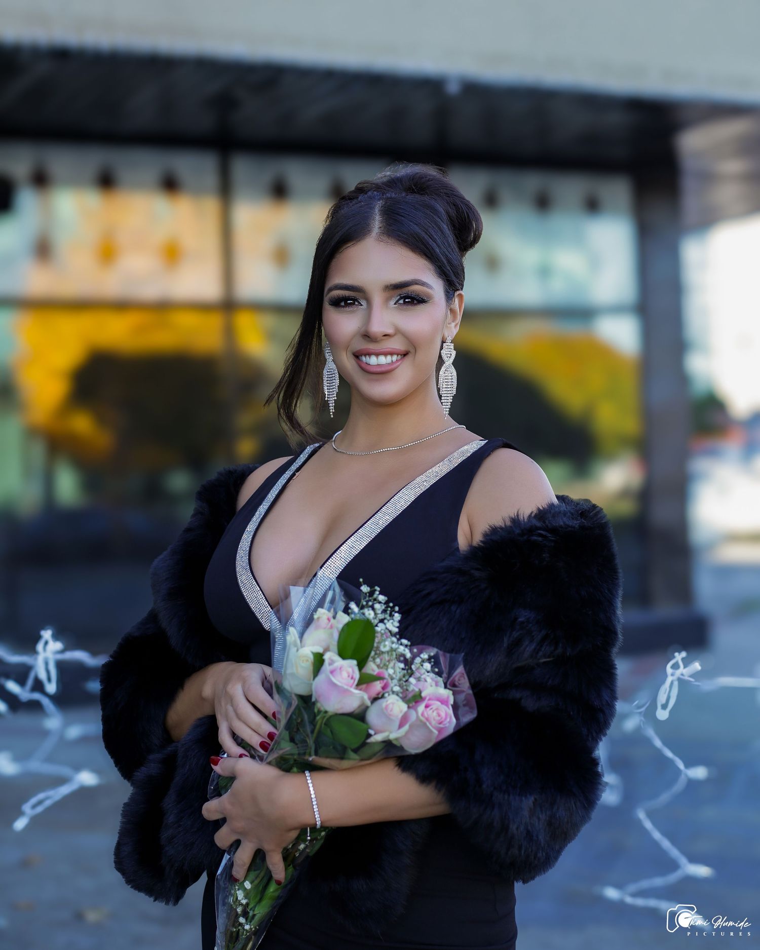 Elegant portrait of someone in a black evening gown holding pink roses outside a building.