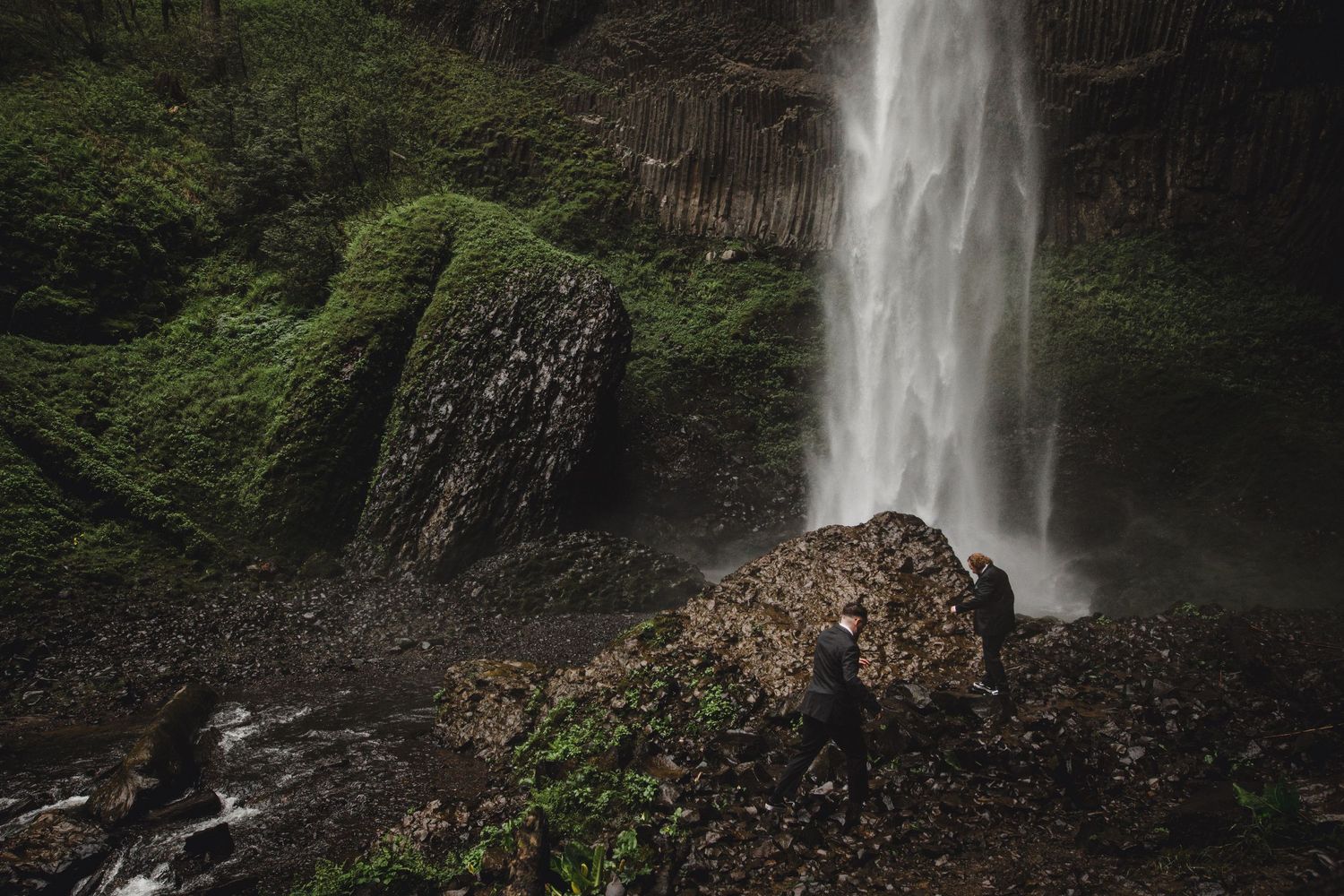 A powerful waterfall cascades down mossy rock formations surrounded by lush green vegetation in a misty forest canyon.