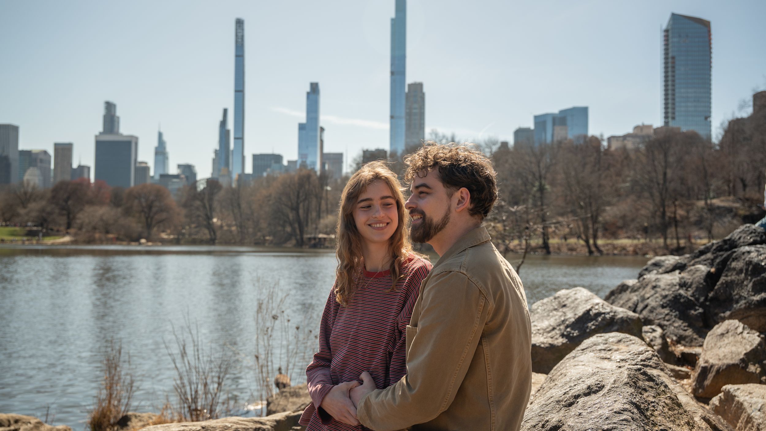 Smiling couple posing together on rocks by Central Park lake with NYC skyline in background.
