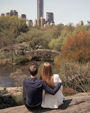 Couple sitting together on a rock overlooking Gapstow Bridge and NYC skyline in Central Park.