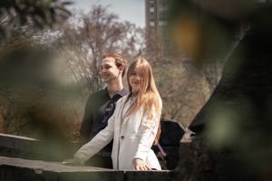 Smiling woman with man standing behind her on a bridge, framed by blurred green leaves in foreground.