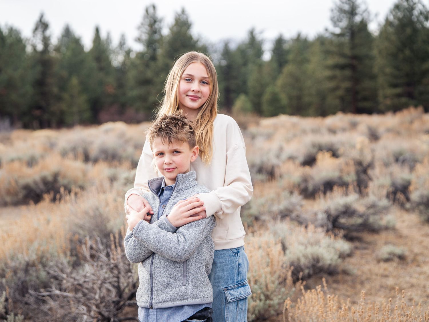A teenage girl hugs a young boy outdoors in a sagebrush field with pine trees in the background.