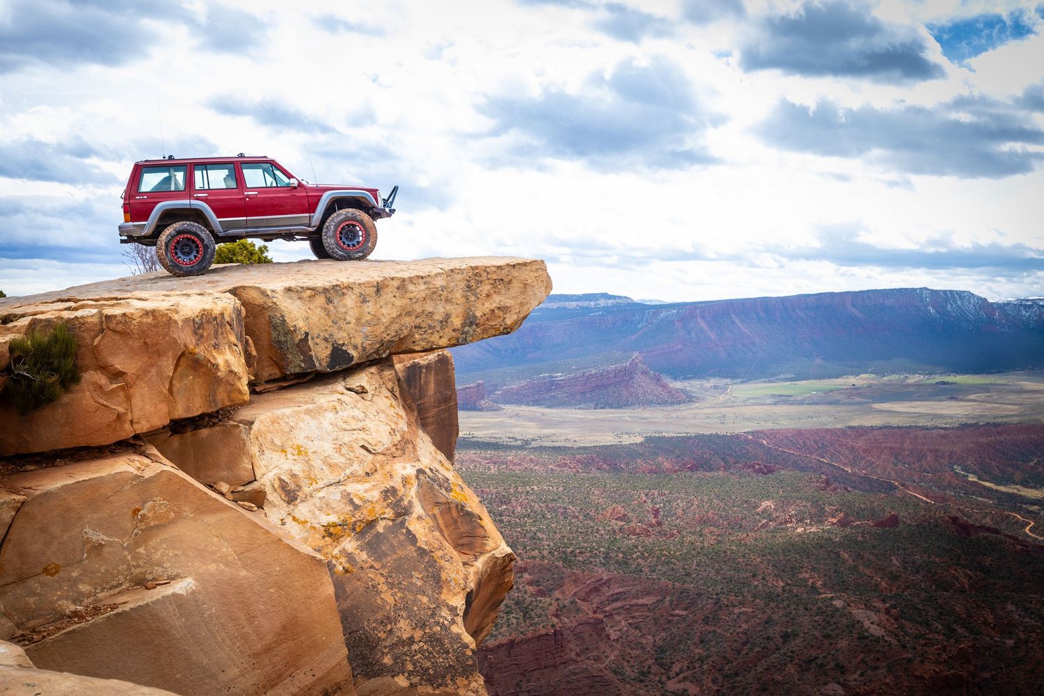 Red SUV parked on a rocky cliff edge overlooking a vast canyon landscape with colorful desert terrain below. Captured by Slate Media (best