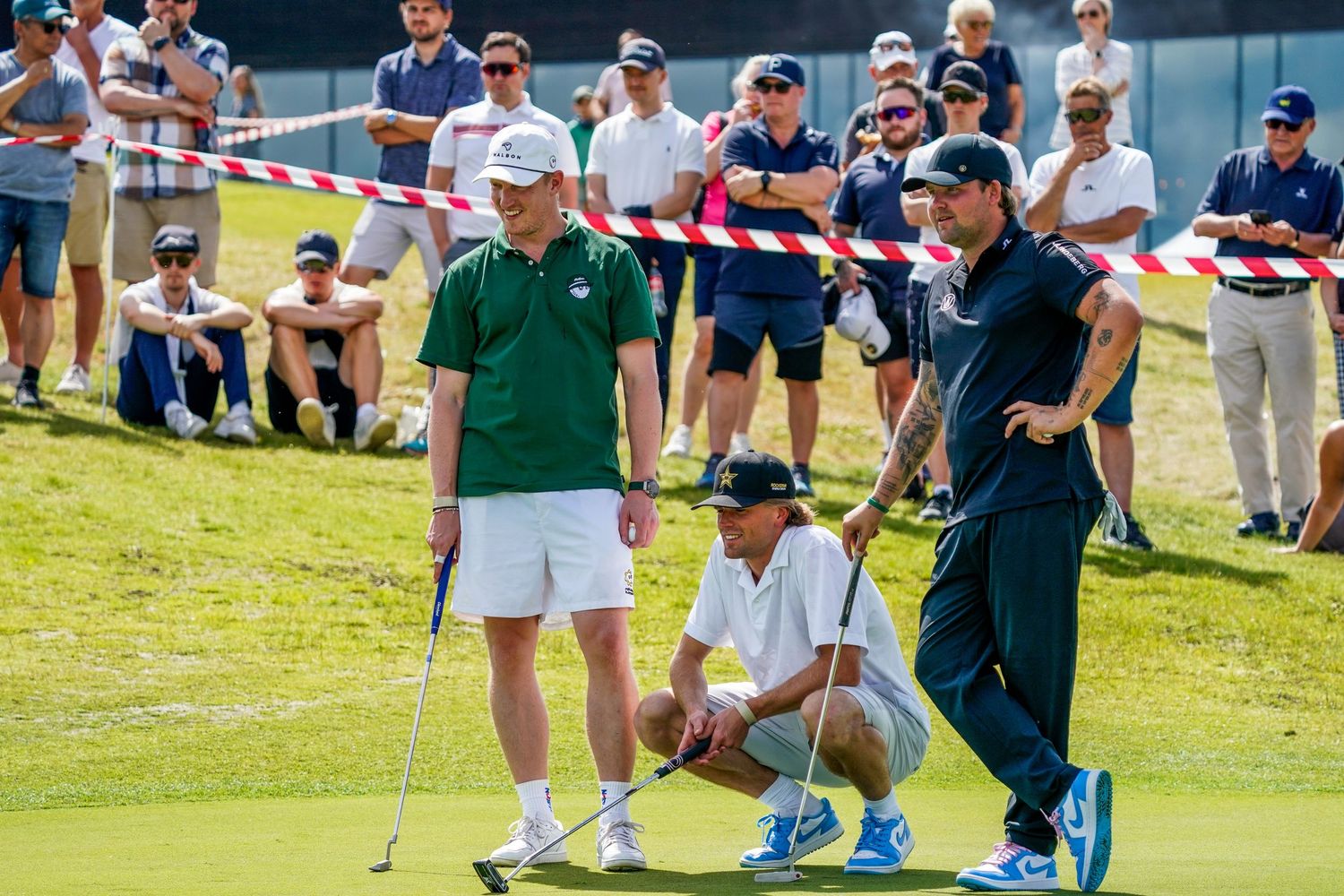 Three golfers on a putting green at a golf tournament, with spectators watching behind rope barriers on a sunny day.