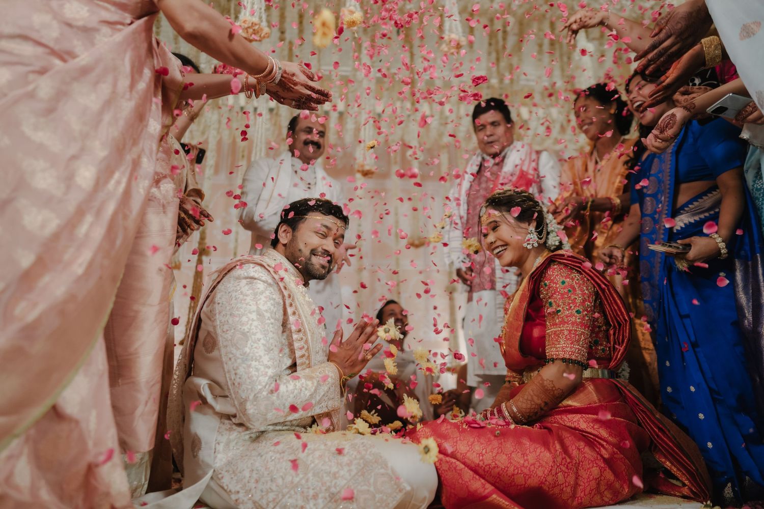 Indian bride and groom smile joyfully as flower petals shower them during traditional Hindu wedding ceremony.