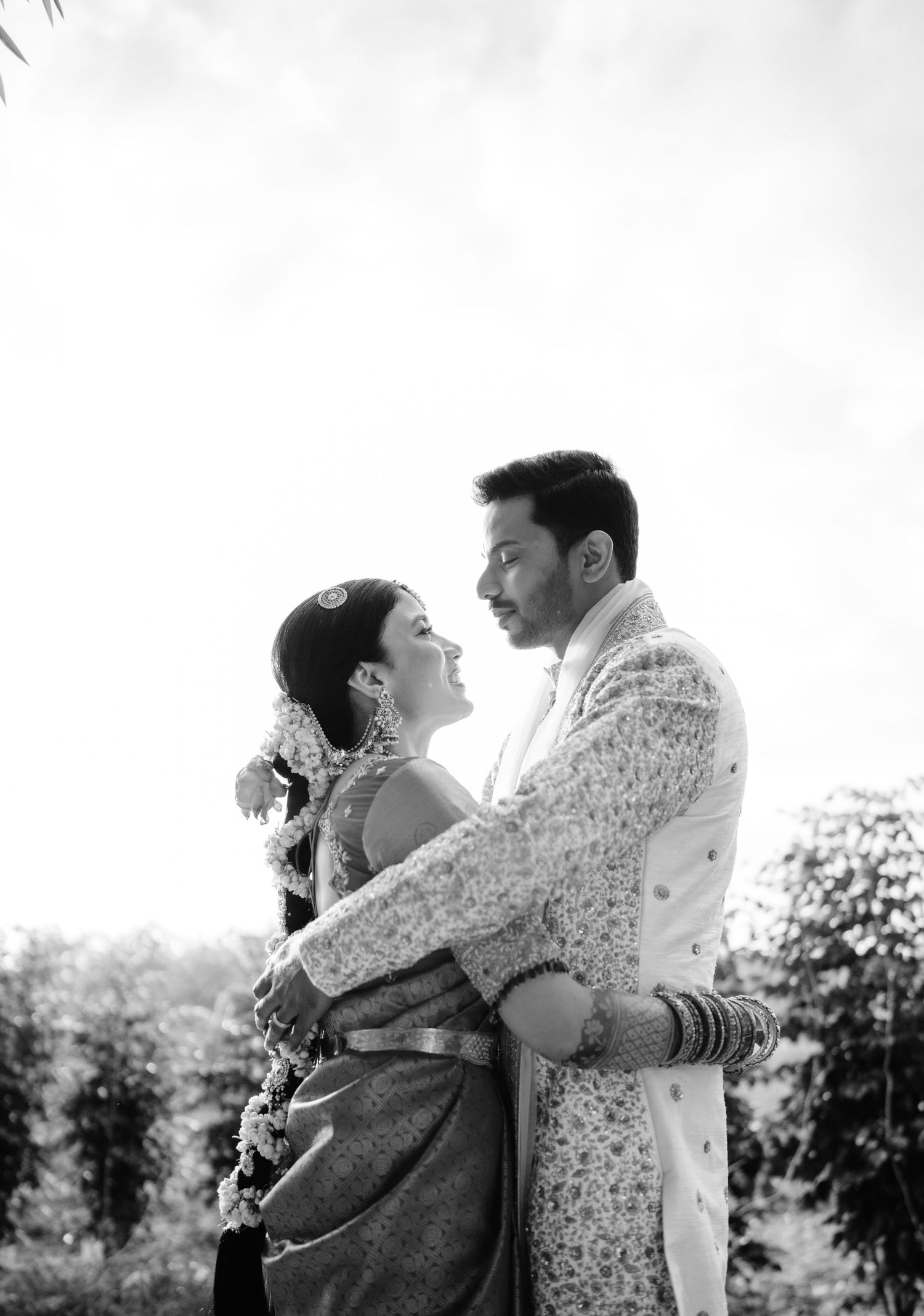 Black and white portrait of smiling Indian couple embracing, bride in saree with jasmine hair garland.