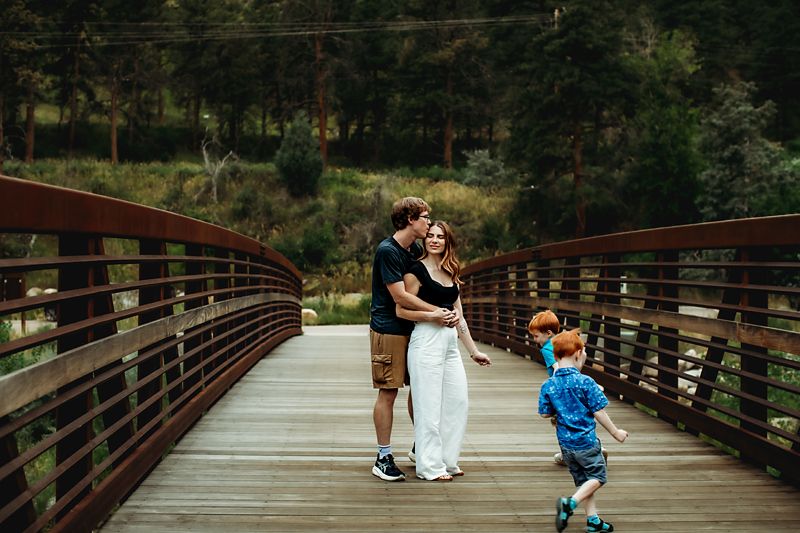 Couple embracing on a wooden bridge in a forest setting while a young red-haired boy runs nearby.