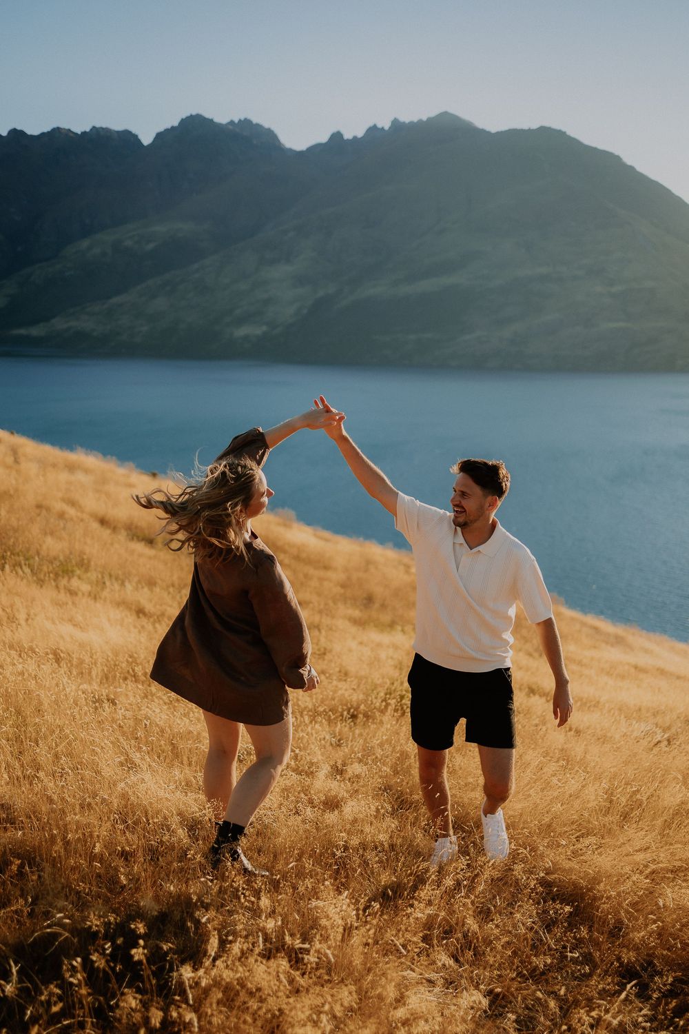 Two people dance playfully in a golden field with mountains and a blue lake in the background.