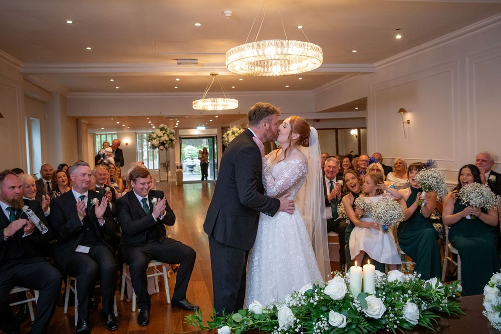 Bride and groom share first kiss at wedding ceremony while guests applaud in elegant chandelier-lit venue.
