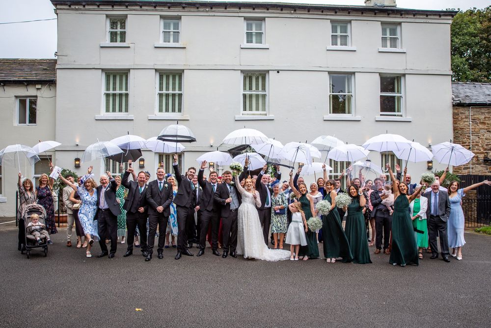 Wedding party and guests celebrate outside historic venue holding white parasols in the air.