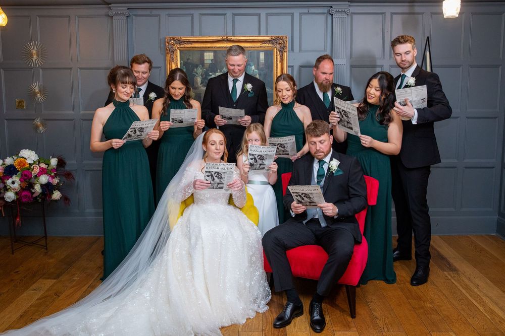 Bridal party in emerald green dresses reads newspapers beside seated bride and groom in stylish venue.
