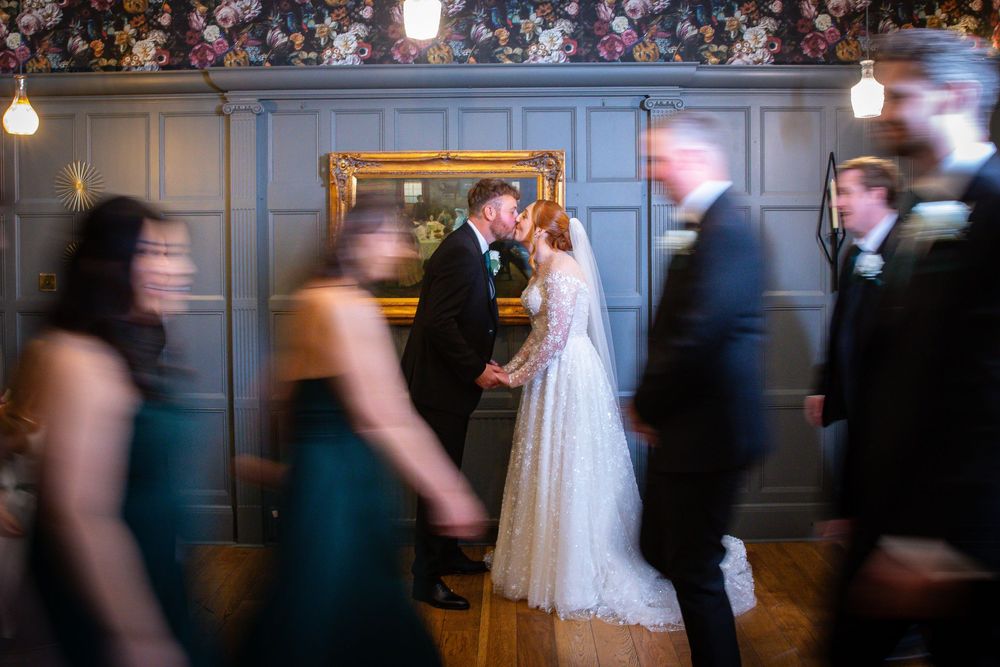 Bride and groom kiss near gold-framed mirror while wedding guests move around them in elegant room.