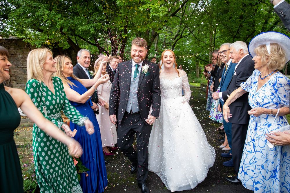 Smiling bride and groom walk through confetti shower surrounded by cheering guests outdoors.