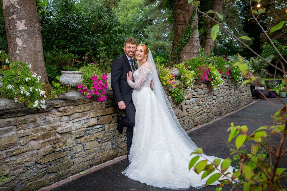 Bride and groom pose together in lace wedding gown beside stone wall with colorful garden flowers.