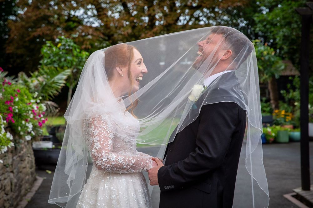 Laughing bride and groom share a tender moment together under flowing bridal veil outdoors.