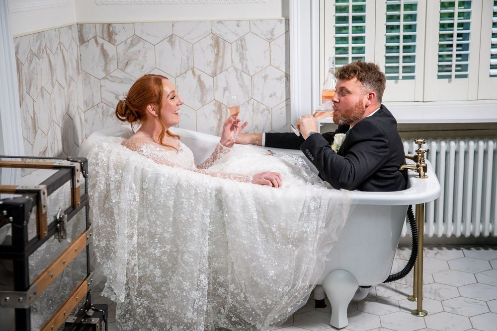 Bride and groom sit playfully in a vintage clawfoot bathtub sharing champagne in elegant bathroom.