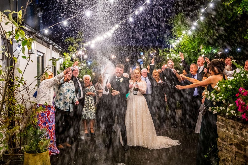 Bride and groom celebrate at night with sparkling confetti cannon surrounded by cheering wedding guests.