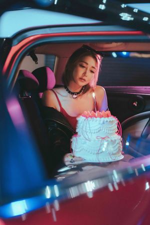 A person sits in a vintage car with a blue and pink decorated birthday cake, illuminated by neon lighting.一個人坐在一輛老式汽車裡，車前放著一個藍粉相間的生日蛋糕，霓虹燈照亮