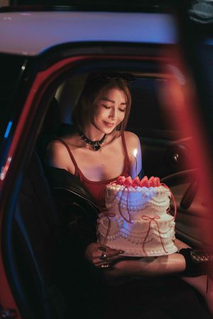 A person holds a birthday cake with glowing candles in a dimly lit car interior illuminated by blue and red lights.一個人手捧生日蛋糕，蛋糕上插著閃閃發光的蠟燭