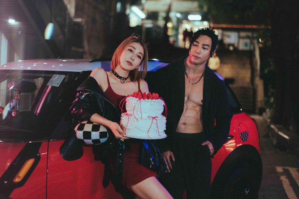 A stylish couple poses at night by a red car, woman holding a decorated cake, wearing edgy fashion outfits.一對時尚情侶在夜色中站在一輛紅色汽車旁擺姿勢拍照