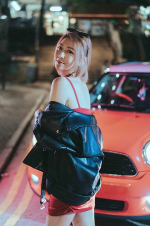 Person in a blue jacket poses next to a red sports car at night with ambient city lighting in the background.夜幕降臨，一名身穿藍色夾克的人站在一輛紅色跑車旁