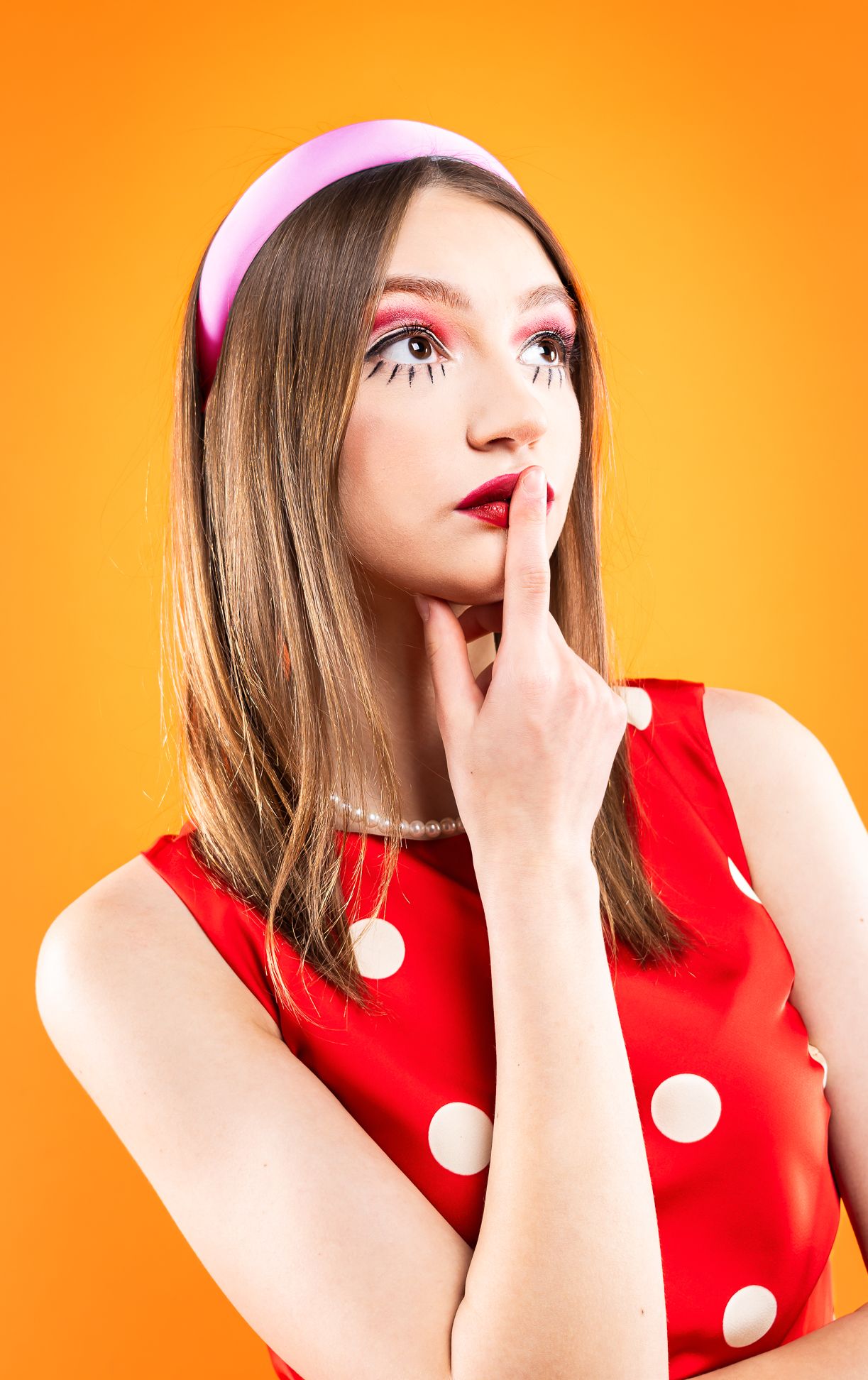 Girl in a red polka dot dress with red dots, a pink headband with a finger on her mouth, looking right and up