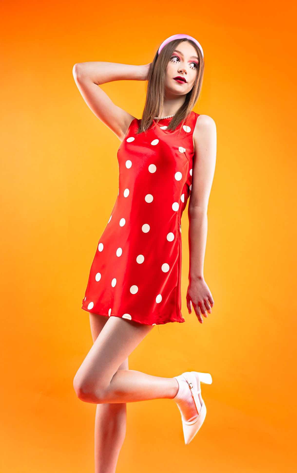 Girl in a red polka dot dress with red dots, a pink headband in front of an orange backdrop with hands on head and leg popped up