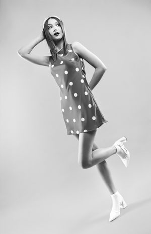 Girl in a red polka dot dress with red dots, a pink headband with hand on head and leg popped up, black and white image