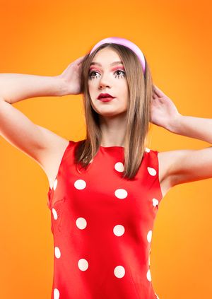 Girl in a red polka dot dress with red dots, a pink headband in front of an orange backdrop with hands on head, looking up and left