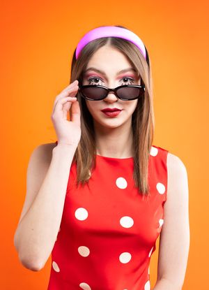 Girl in a red polka dot dress with red dots, a pink headband in front of an orange backdrop with black sunglasses, pointing them down