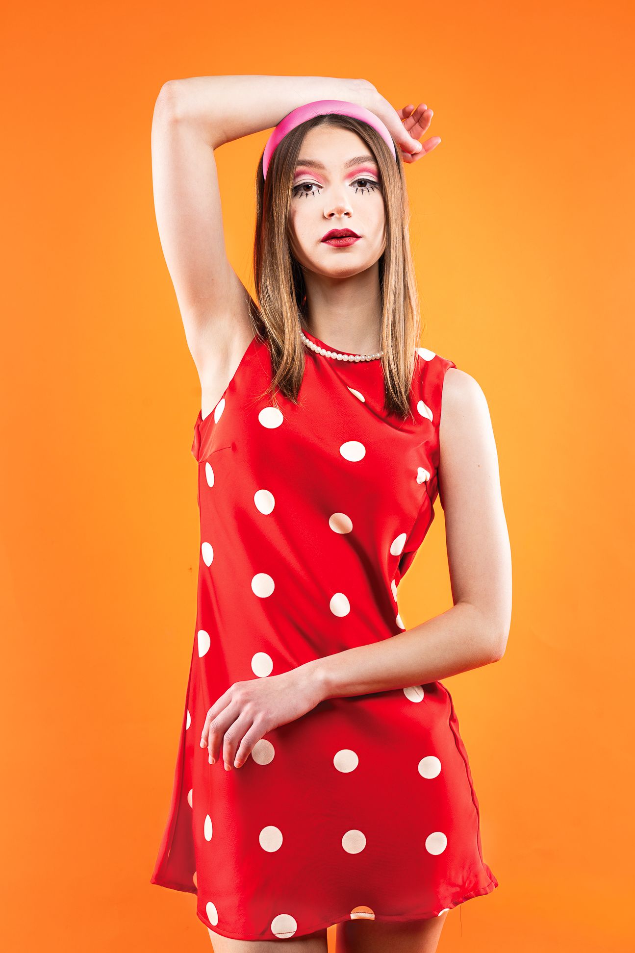 Girl in a red polka dot dress with red dots, a pink headband in front of an orange backdrop with hands on top of head and across body