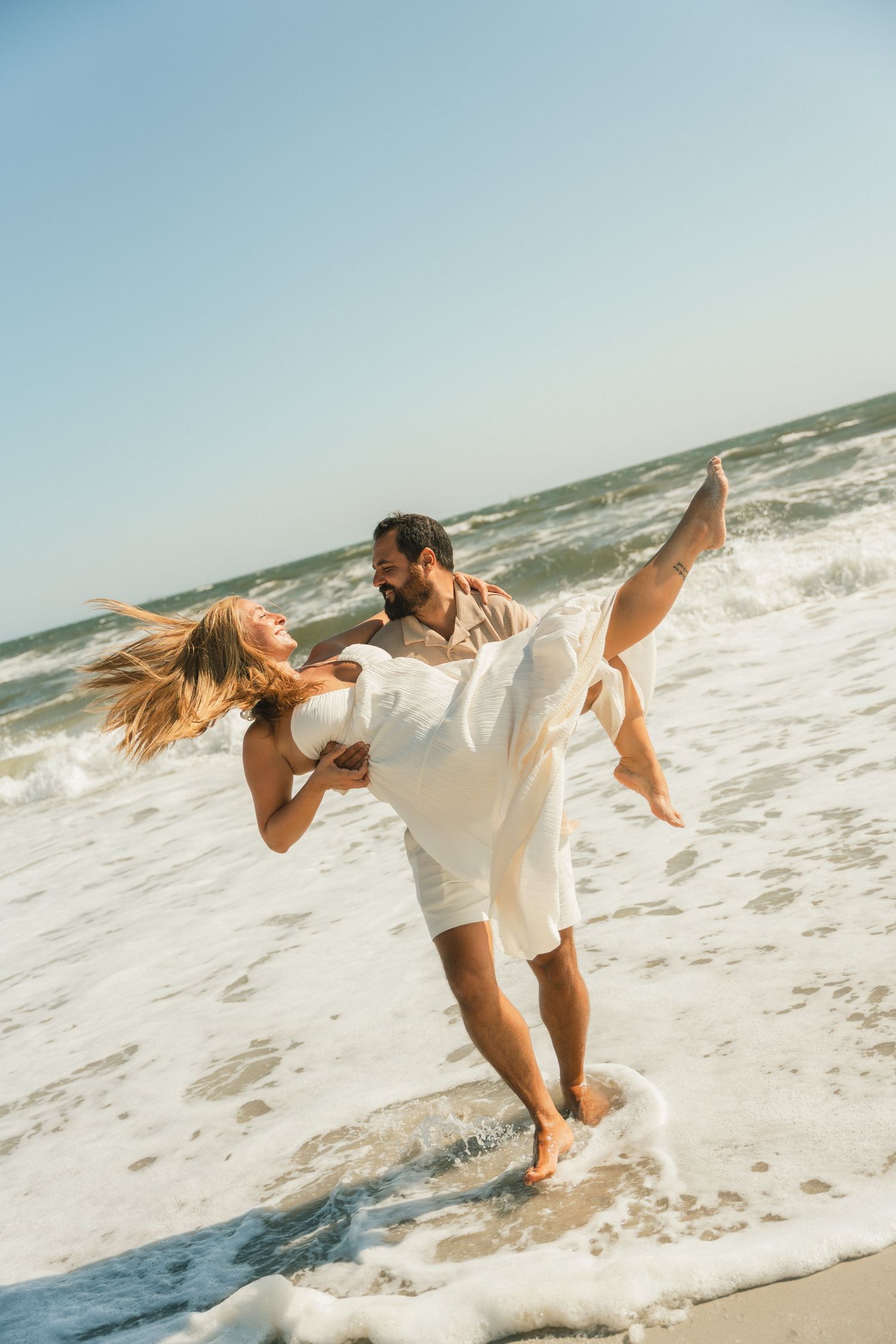 Couple plays in ocean waves wearing white outfits during beach photoshoot.