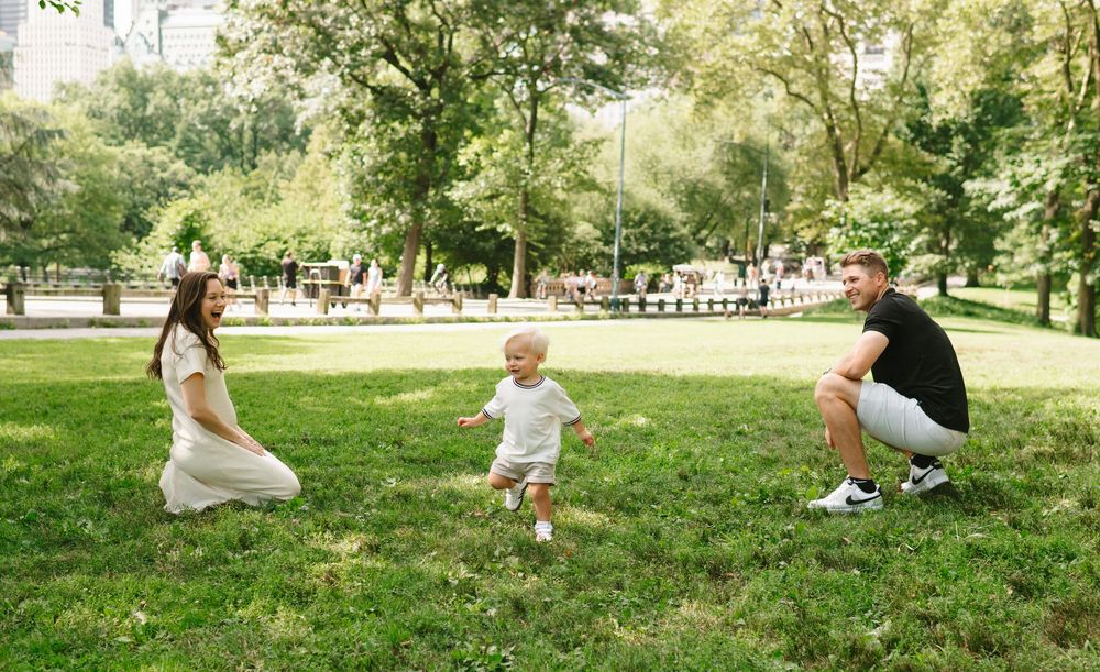 A toddler takes steps on grass in a park while two adults crouch on either side during a sunny day.