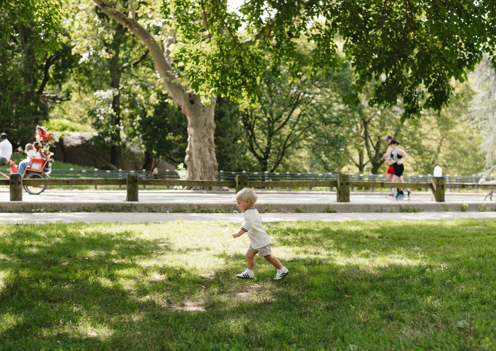 A small child in white clothing runs across a sunny park lawn with trees and people in the background.