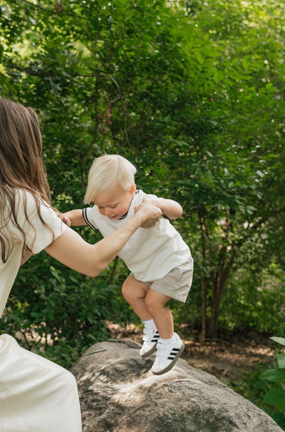 A toddler in white clothing and striped socks explores a large rock in a lush garden setting while being supported.