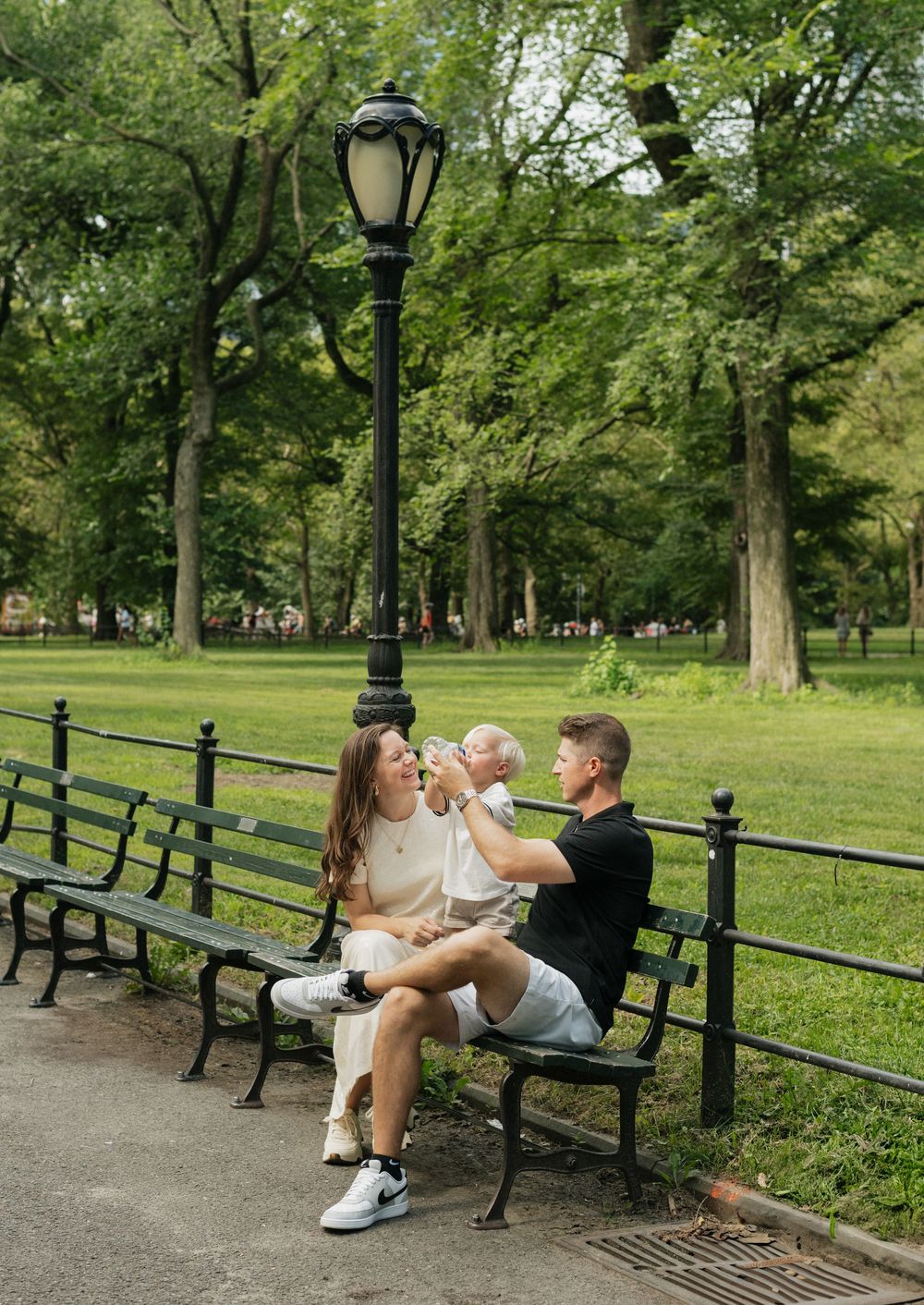 A couple sits on a park bench with their small dog near a vintage lamp post in Central Park on a sunny day.