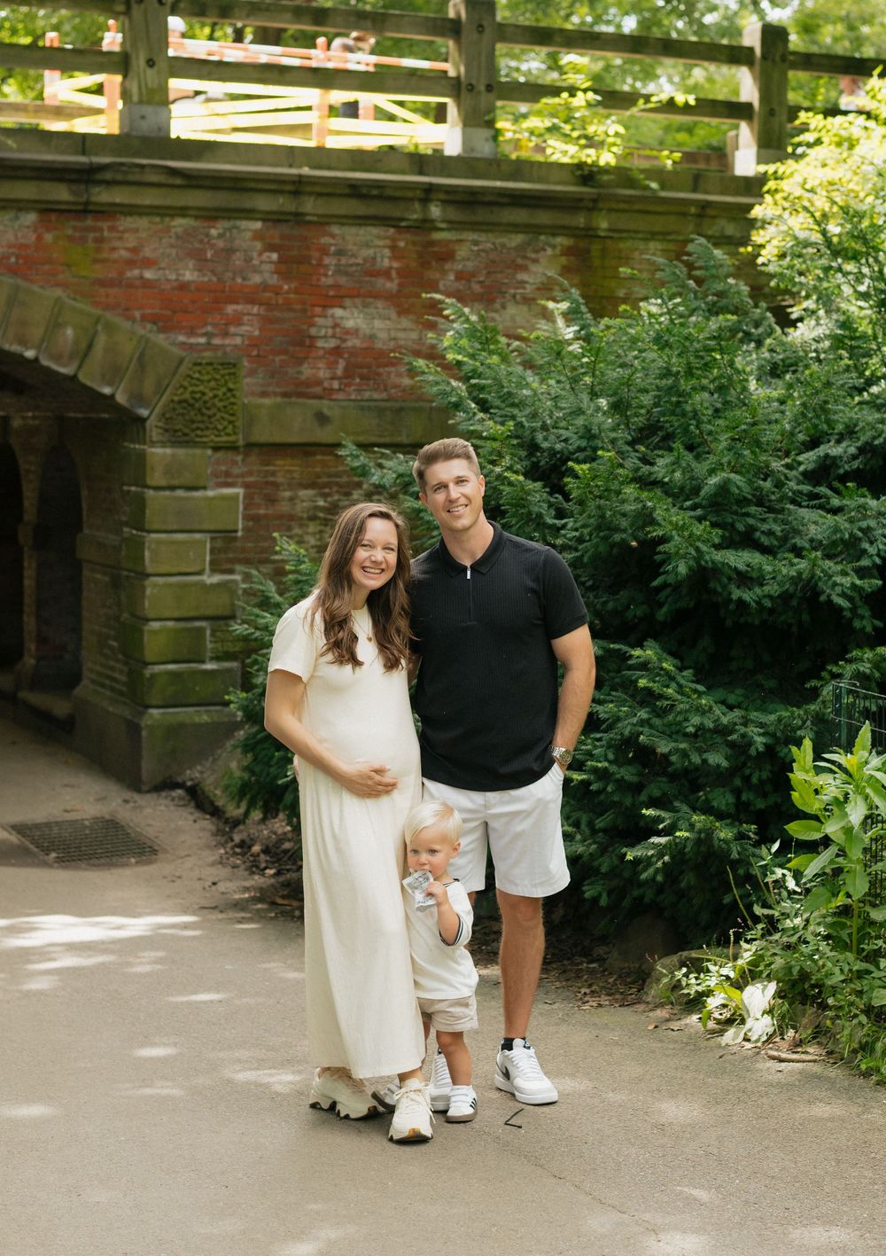 A family poses together on a path under a stone bridge in a lush garden setting during summer.
