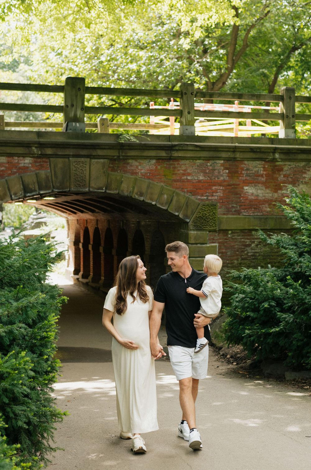 A family walks along a shaded path under a brick bridge in a lush park setting on a sunny summer day.