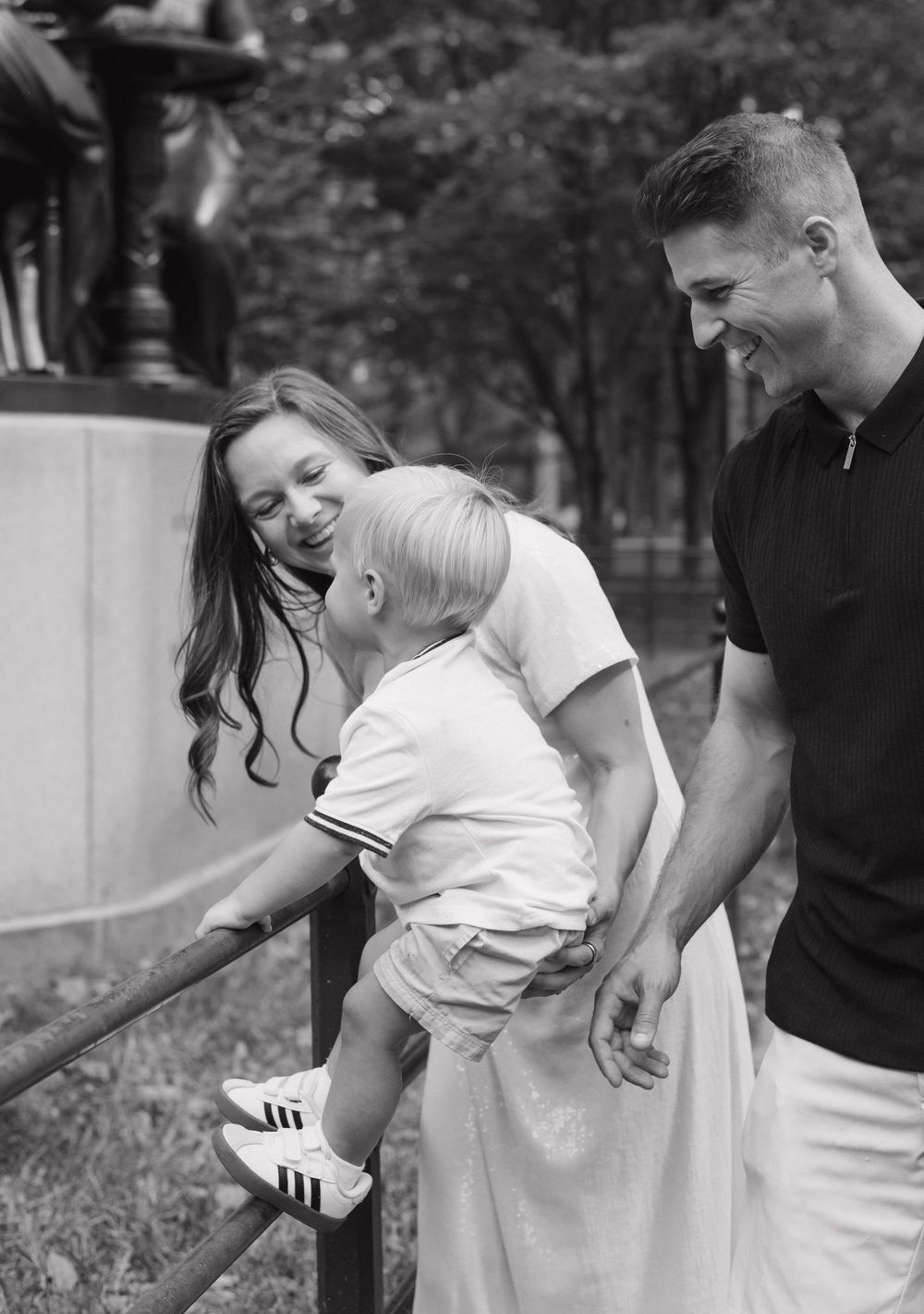 A candid black and white family moment shows parents laughing together with their young child outdoors.