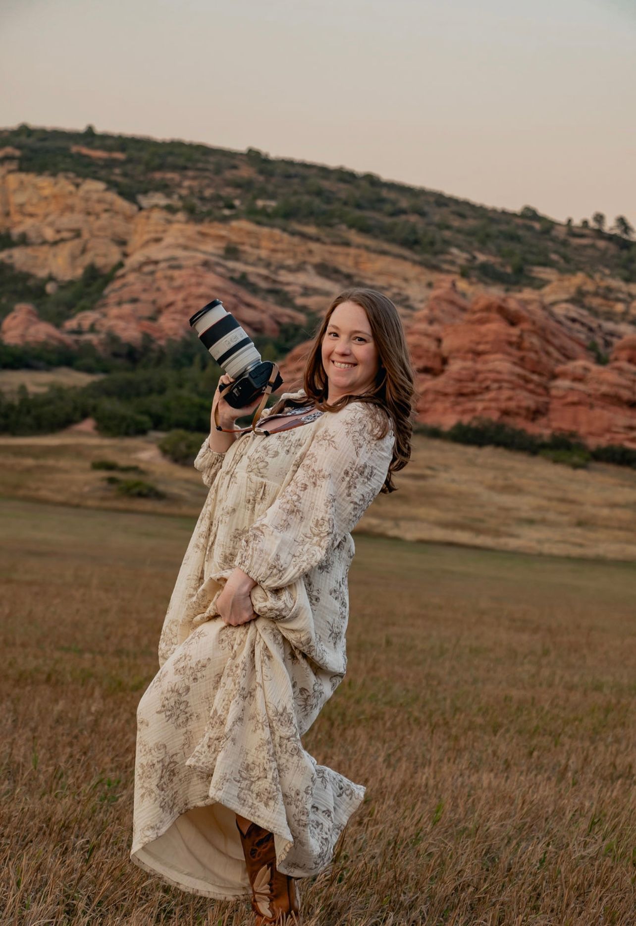 Professional photographer in flowing cream lace dress poses with camera against backdrop of red rock formations at sunset.