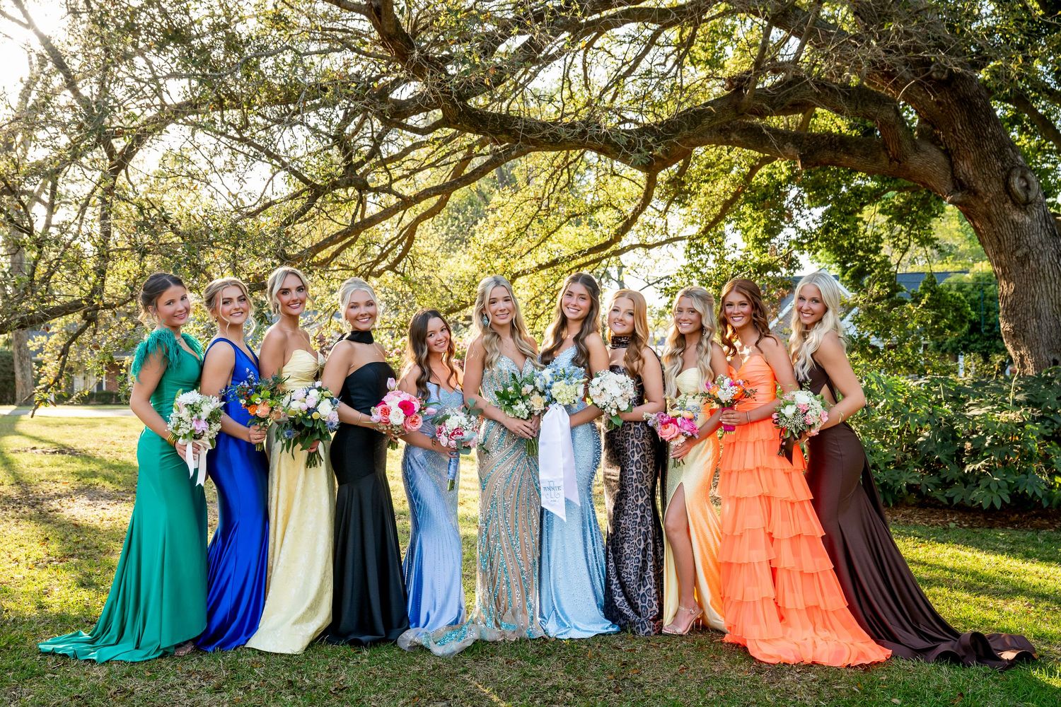 Bridesmaids in colorful mismatched gowns pose outdoors under a large oak tree holding floral bouquets.