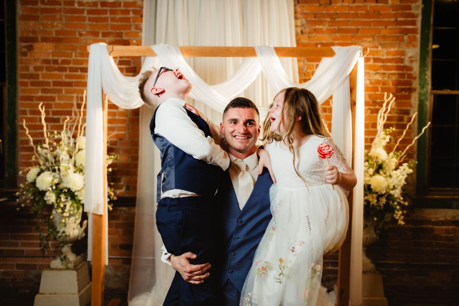 Bride and groom celebrate with young child at rustic brick wedding venue with floral arrangements and draped white fabric backdrop.
