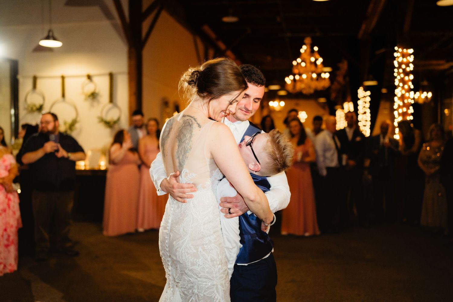 Bride and groom share emotional first dance embrace at rustic wedding reception with warm chandelier lighting.