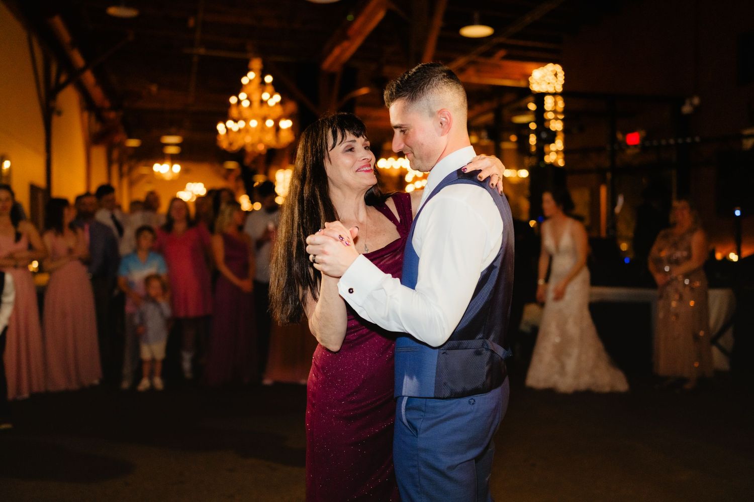 A couple shares a romantic first dance at a wedding reception in a warmly lit venue with guests watching in the background.